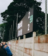 A large digital scoreboard at an outdoor sports venue displays a score of 2-2. Spectators are gathered, observing the event with interest. The scene is enclosed by a tall metal fence and surrounded by lush green trees, indicating a natural setting. Overcast skies add a muted tone to the environment.