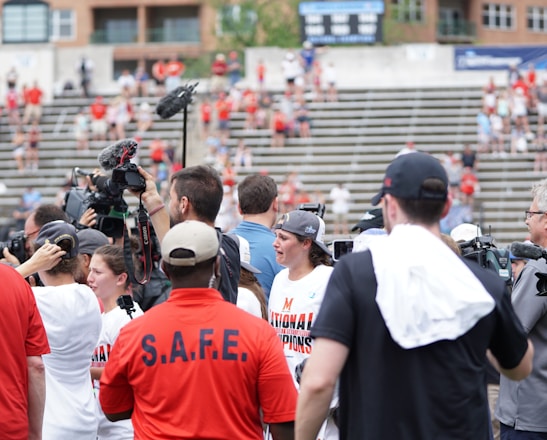 A lively gathering in an outdoor sports venue with a group of people, some wearing matching shirts labeled 'National Champions.' Several individuals are holding cameras and microphones, indicating a media presence. The background shows blurred spectators sitting on bleachers.