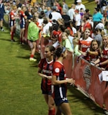 The image features a lively gathering of young soccer players and fans next to a field. Several female athletes in red and blue uniforms interact with an enthusiastic crowd standing behind a barrier. Some people in the crowd are holding objects, like pens and paper, possibly for autographs.
