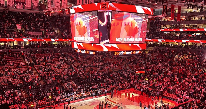 An indoor sports arena filled with spectators and a basketball court illuminated by red lighting. The scoreboards and video screens display promotional content, with numerous banners and championship flags hanging from the ceiling. Players and officials are gathered on the court, possibly in preparation for a game or event.
