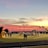 A sports field with a football game unfolding under a vibrant sunset sky, featuring a large inflatable mascot and a scoreboard displaying the score. Players and coaches are visible on the green field, surrounded by a track and some flags waving in the breeze.