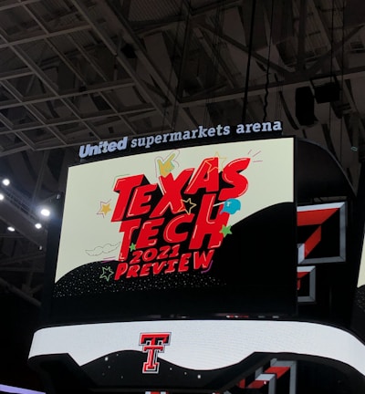 A scoreboard displaying the text 'Texas Tech 2021 Preview' in bold red letters with decorative elements like stars and clouds. The background is predominantly dark with bright accent colors, and the setting is within an indoor sports arena, specifically the United Supermarkets Arena.