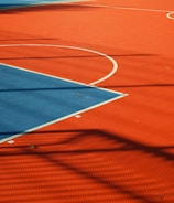 A vibrant sports court with sections of red and blue, featuring crisp white lines marking the boundaries. Shadows from a structure create geometric patterns across the surface, adding depth and interest to the scene.